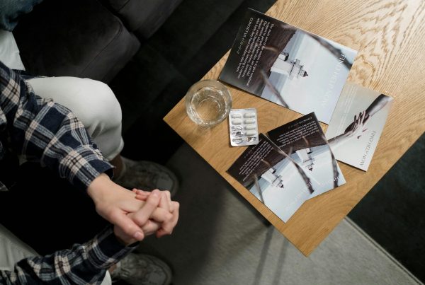 Person sitting by table with pills, glass of water, and brochures, symbolizing persistent symptoms linked to hormones, gut health, toxins, stress, and the need for root cause healing with the Root Cause Profile Quiz.